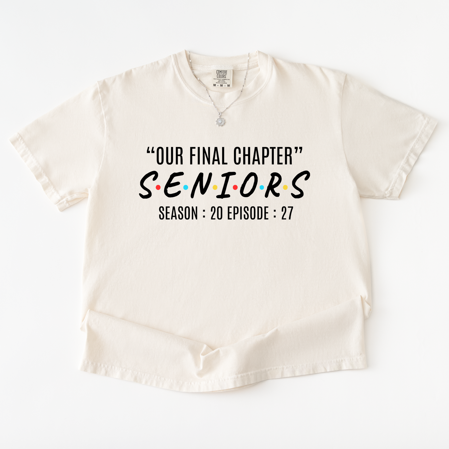 An off-white short-sleeve t-shirt with a graphic print on the front. The print features the text '“OUR FINAL CHAPTER” SENIORS SEASON : 20 EPISODE : 27' styled like the iconic Friends TV show logo, with the word 'SENIORS' in black and the letter accents in red, yellow, and blue.