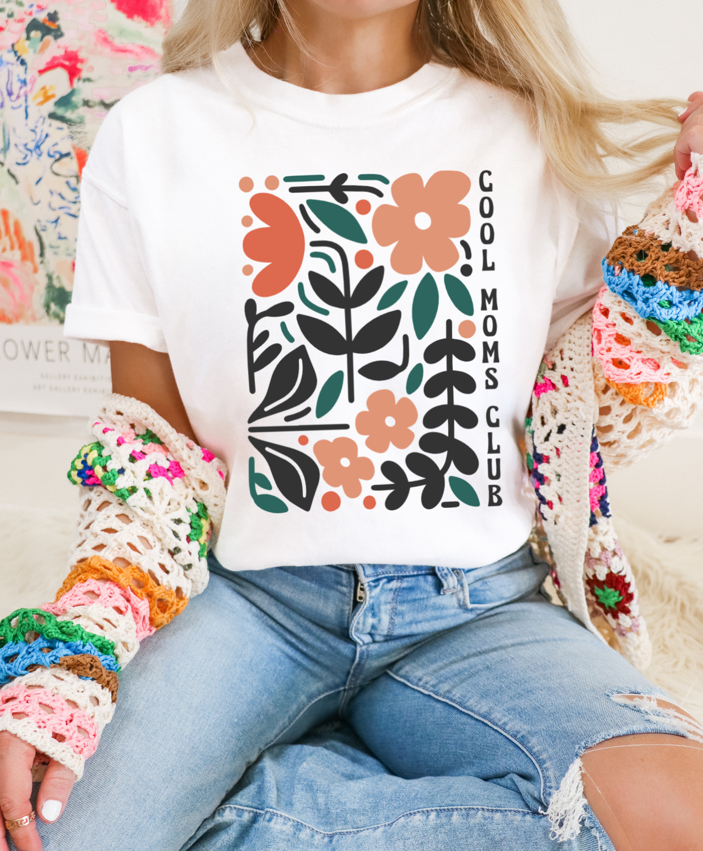 Person wearing a white t-shirt with floral design and 'Cool Moms Club' text, sitting against a colorful background.