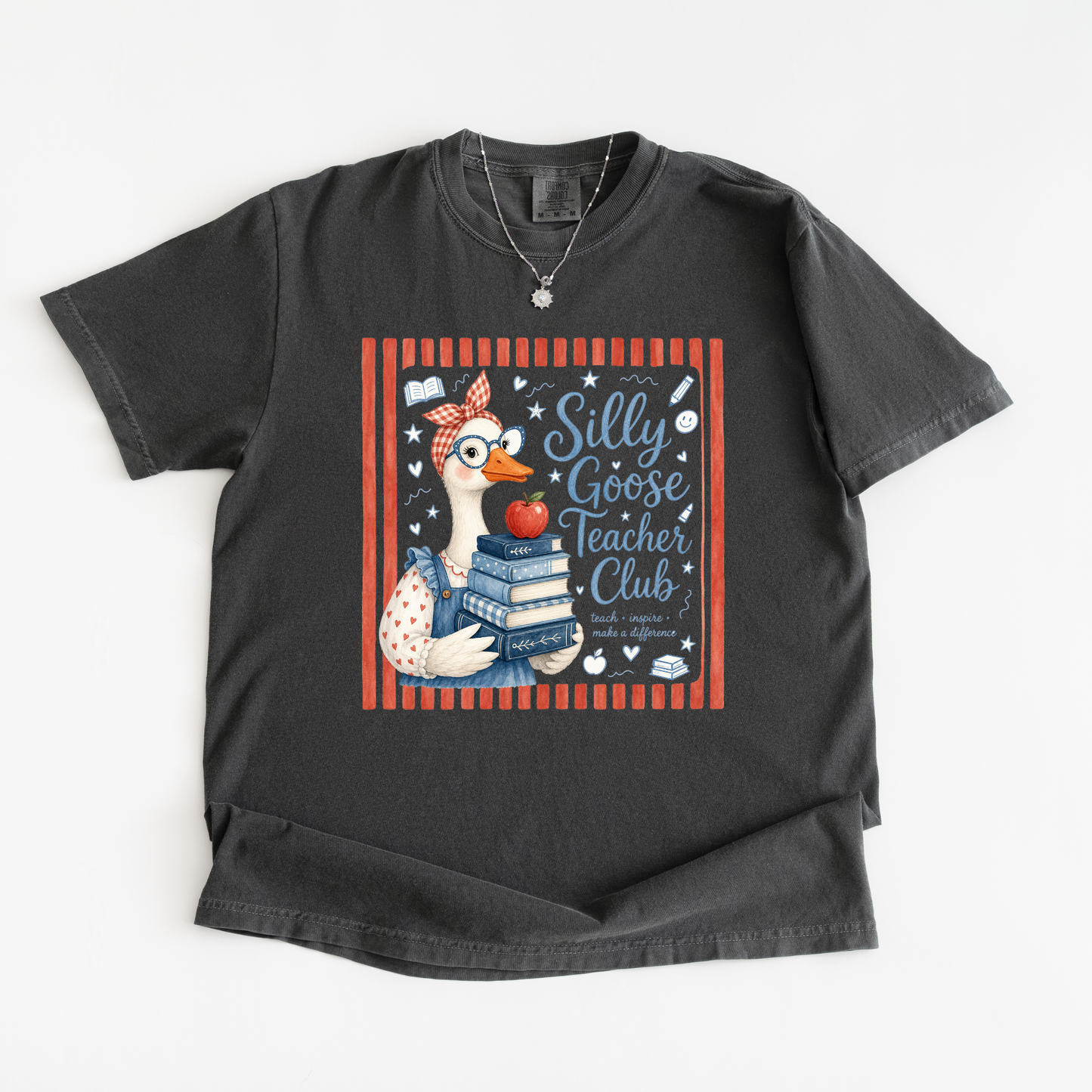 A dark charcoal gray t-shirt with a graphic print of a goose wearing glasses and a headband, holding books and an apple, with the text 'Silly Goose Teacher Club' inside a red striped border.