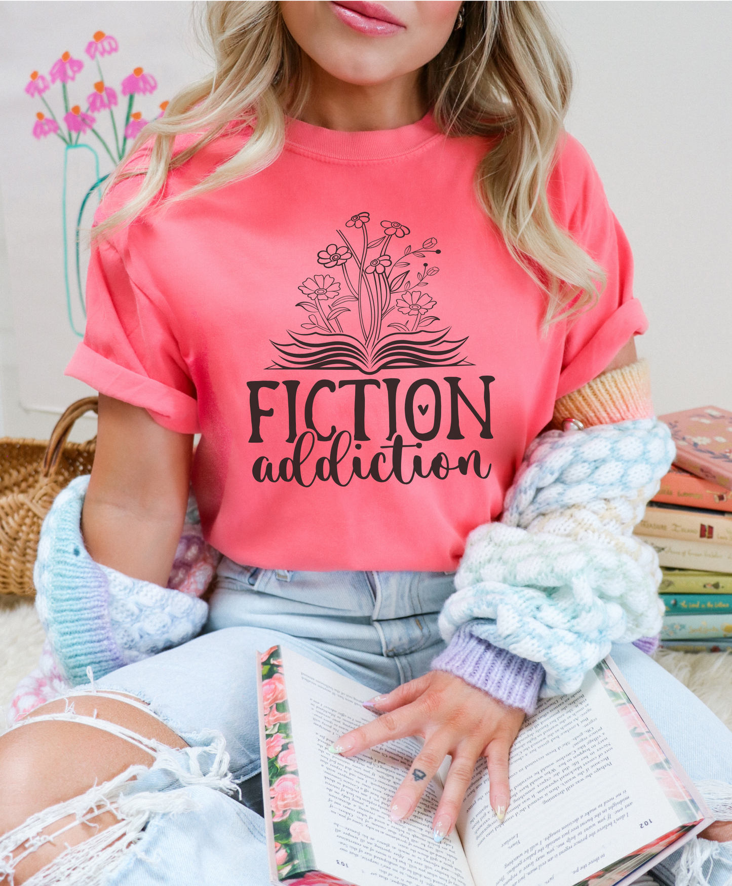 Person wearing a pink t-shirt with 'Fiction Addiction' design, holding a book.