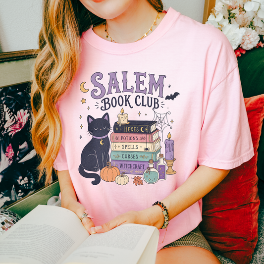 A woman wearing a light pink graphic t-shirt with a 'Salem Book Club' witchcraft illustration, holding an open book in a cozy indoor setting.