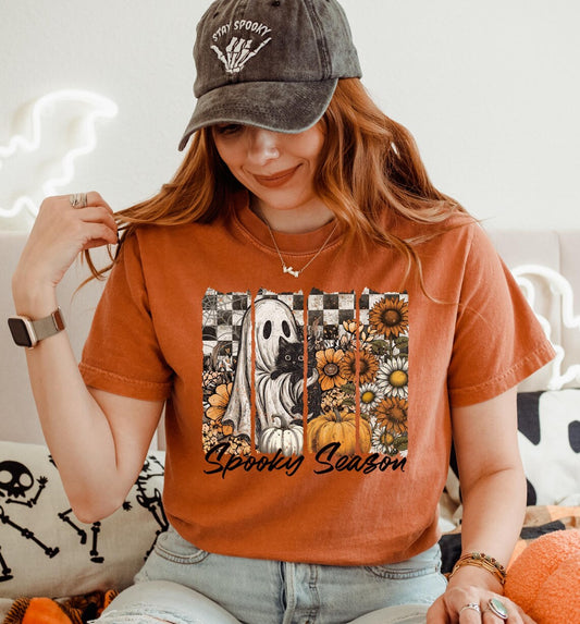 A woman wearing a burnt orange graphic t-shirt with a Halloween design featuring a ghost, pumpkins, sunflowers, and the text 'Spooky Season'. She is also wearing a dark gray baseball cap with 'ITY EPDROY' embroidered on it, sitting in a cozy indoor setting with Halloween-themed pillows.