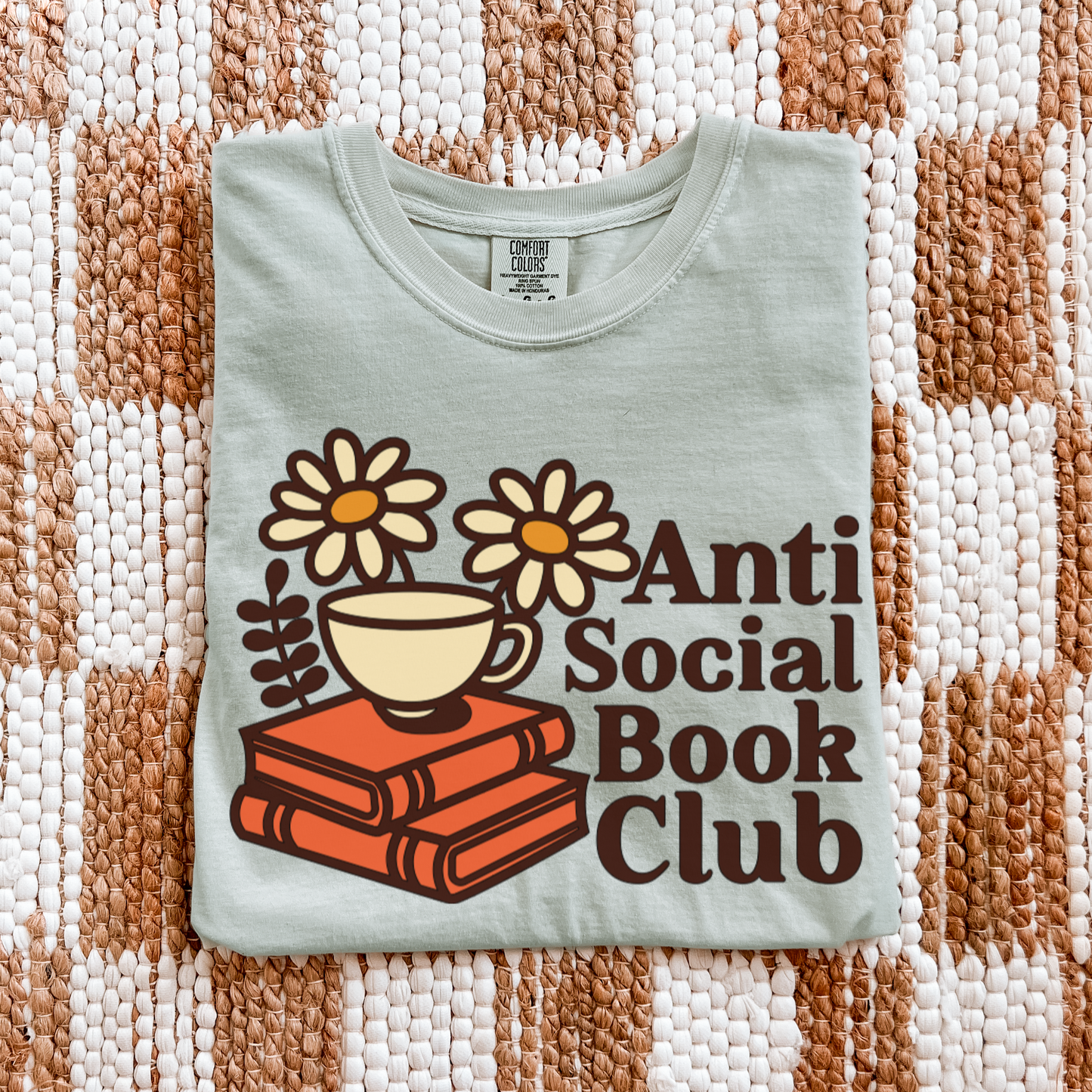 Light green t-shirt with 'Anti Social Book Club' design featuring a cup, books, and flowers on a textured fabric background.