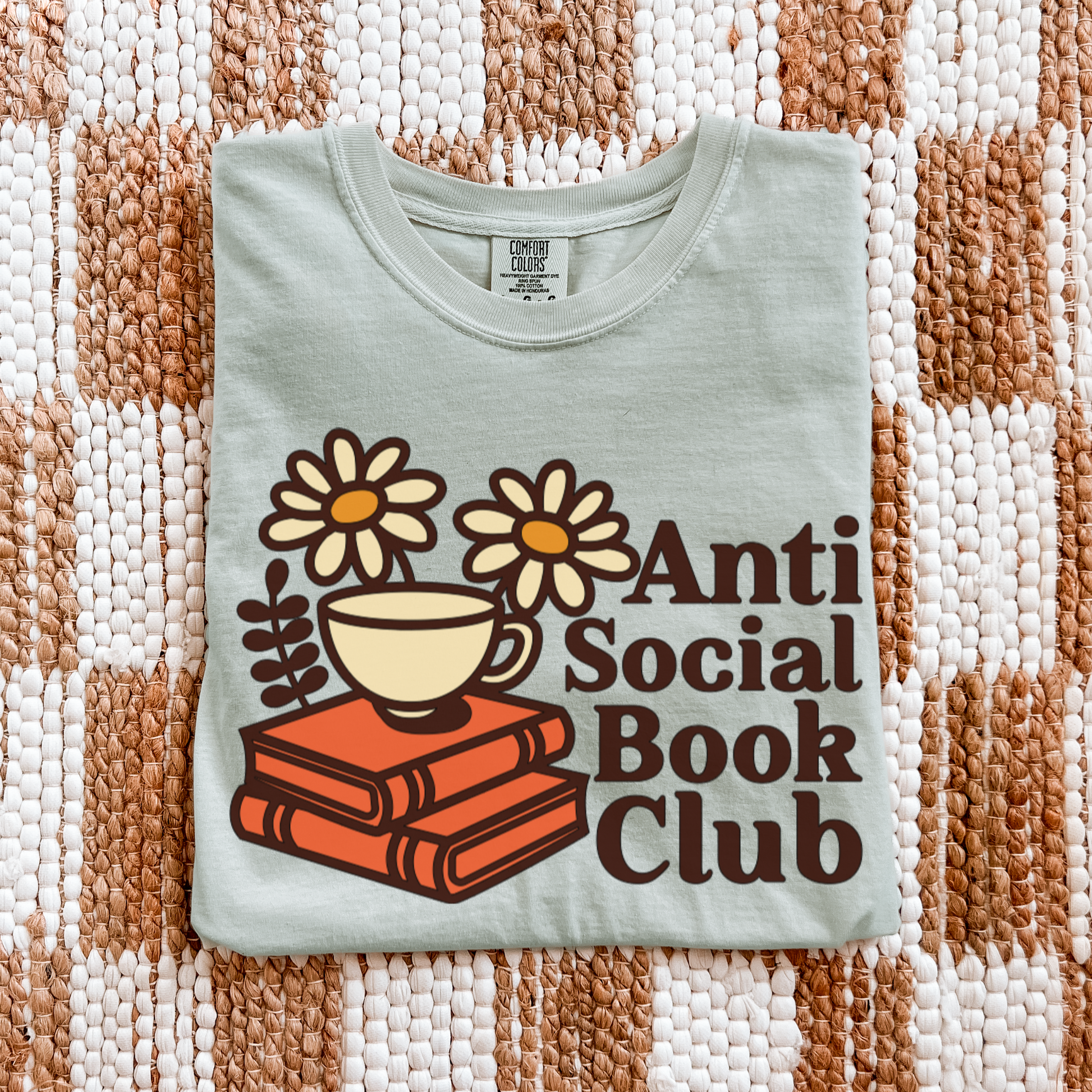 Light green t-shirt with 'Anti Social Book Club' design featuring a cup, books, and flowers on a textured fabric background.