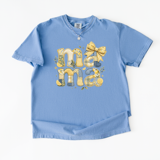 A light blue short-sleeve t-shirt with a decorative graphic spelling 'mama' in yellow and white plaid socks, decorated with a yellow plaid bow and small floral details, shown on a white background.