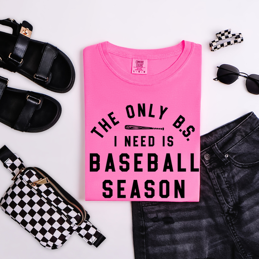 Pink t-shirt with 'The Only BS I Need is Baseball Season' text, surrounded by black sandals, sunglasses, and a checkered bag on a white background.