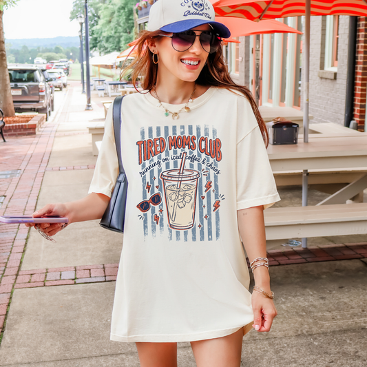 A woman wearing an oversized cream-colored graphic t-shirt with a 'Tired Moms Club' drink design, styled with a blue cap, sunglasses, and a shoulder bag, standing on an outdoor street.